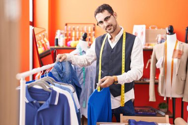 Young hispanic man tailor smiling confident holding clothes on rack at atelier