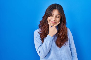 Hispanic young woman standing over blue background looking confident at the camera smiling with crossed arms and hand raised on chin. thinking positive. 
