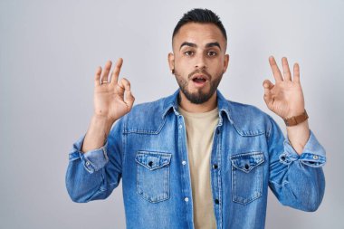 Young hispanic man standing over isolated background looking surprised and shocked doing ok approval symbol with fingers. crazy expression 
