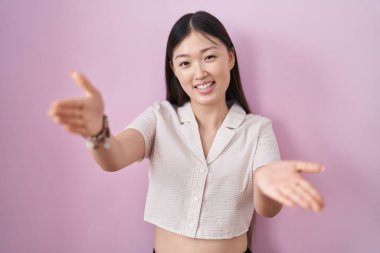 Çinli genç bir kadın pembe arka planda durup kameraya bakarak sarılmak için kollarını açmış gülümsüyor. Mutluluğu kucaklayan neşeli bir ifade.. 