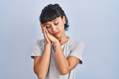 Mavi arka plan üzerine gündelik tişört giyen genç İspanyol kadın yorgun uyuyor ve elleri ile birlikte poz verirken gözleri kapalı gülümsüyor.. 