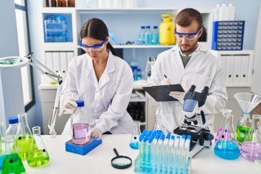 Man and woman scientist partners write on clipboard measuring liquid working at laboratory