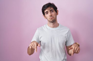 Young hispanic man standing over pink background pointing down looking sad and upset, indicating direction with fingers, unhappy and depressed. 