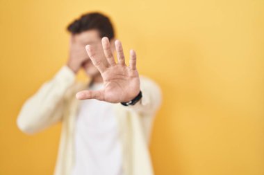 Sarı arka planda dikilen genç İspanyol adam elleriyle gözlerini kapatıyor ve üzgün ve korku ifadeleriyle jestleri durduruyor. utangaç ve olumsuz kavram. 