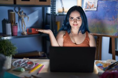Young modern girl with blue hair sitting at art studio with laptop at night smiling cheerful presenting and pointing with palm of hand looking at the camera. 