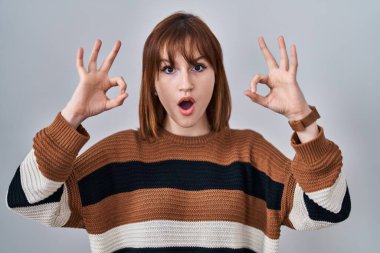 İzole edilmiş arka planda çizgili süveter giyen genç ve güzel bir kadın. Şaşırmış ve şaşırmış görünüyor. Parmaklarıyla onay sembolü yapıyor. çılgın ifade 