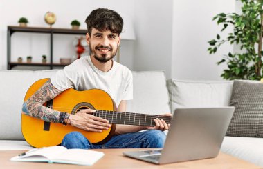 Genç İspanyol adam klasik gitar çalıyor ve evde dizüstü bilgisayar kullanıyor..