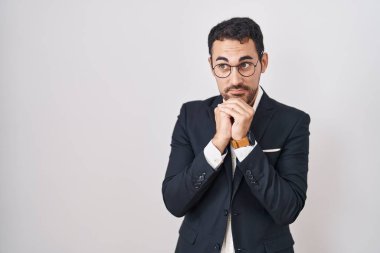 Handsome business hispanic man standing over white background laughing nervous and excited with hands on chin looking to the side 