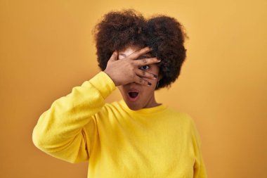 Genç Afrikalı Amerikalı kadın sarı arka planda dikiliyor. Şoka giriyor. Yüzü ve gözleri kapanıyor, utangaç ifadesiyle parmaklarından bakıyor.. 