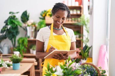 Middle age african american woman florist smiling confident writing on notebook at flower shop