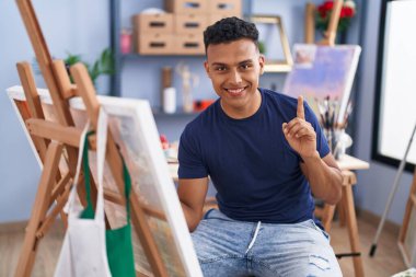 Young hispanic man painting sitting at art studio surprised with an idea or question pointing finger with happy face, number one 