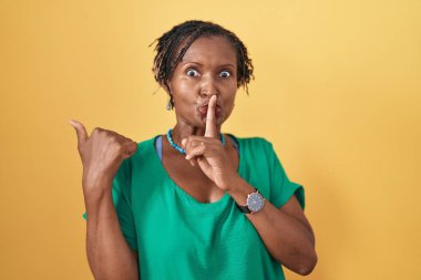 African woman with dreadlocks standing over yellow background asking to be quiet with finger on lips pointing with hand to the side. silence and secret concept. 