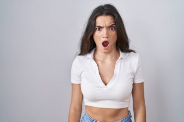 Young teenager girl standing over white background in shock face, looking skeptical and sarcastic, surprised with open mouth 