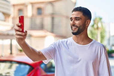 Genç Arap adam kendinden emin gülümsüyor. Sokakta akıllı telefonun yanında selfie çekiyor.