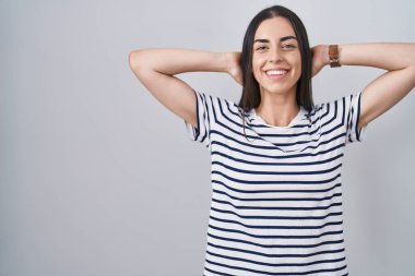 Genç esmer kadın çizgili tişört giyiyor rahatlıyor ve esniyor, kolları ve elleri başının arkasında mutlu bir şekilde gülümsüyor. 