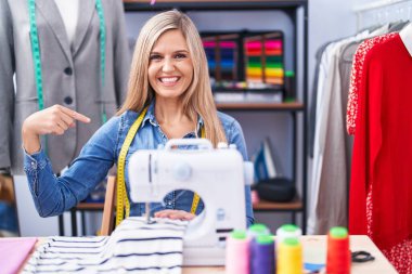 Blonde woman dressmaker designer using sew machine looking confident with smile on face, pointing oneself with fingers proud and happy. 
