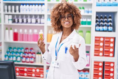 African american woman pharmacist smiling confident holding pills bottles at pharmacy