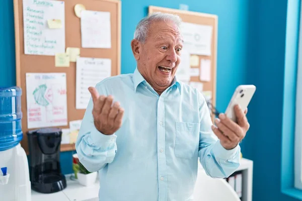 Senior man with grey hair working at the office doing video call with smartphone celebrating achievement with happy smile and winner expression with raised hand