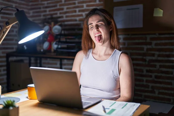Brunette woman working at the office at night sticking tongue out happy with funny expression. emotion concept.
