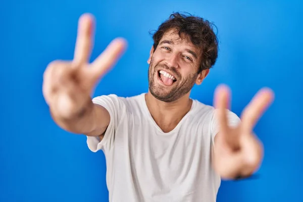 両手の指が勝利記号を行うことを示す舌で笑顔青い背景の上に立ってヒスパニック系の若い男 — ストック写真