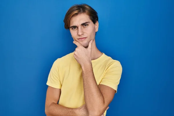 Young man standing over blue background looking confident at the camera with smile with crossed arms and hand raised on chin. thinking positive.