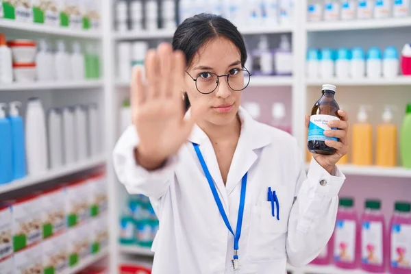 Young hispanic woman working at pharmacy drugstore holding syrup with open hand doing stop sign with serious and confident expression, defense gesture