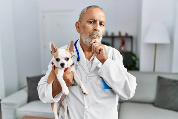 Mature veterinarian man checking dog health serious face thinking about question with hand on chin, thoughtful about confusing idea
