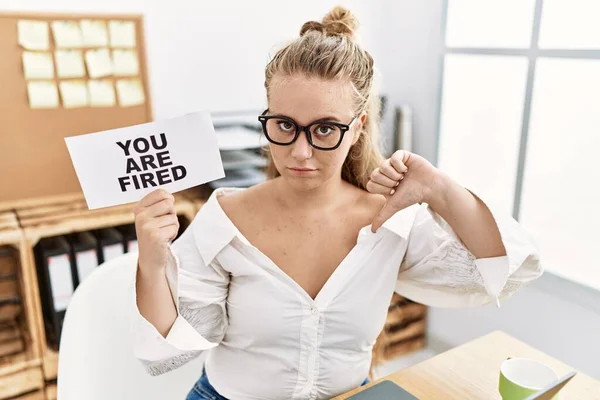 Young caucasian woman holding you are fired banner at the office with angry face, negative sign showing dislike with thumbs down, rejection concept