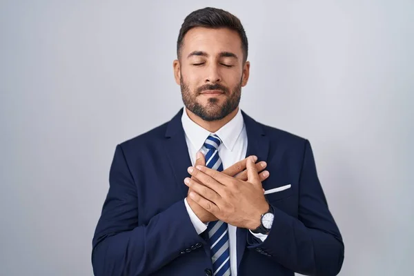 Handsome hispanic man wearing suit and tie smiling with hands on chest with closed eyes and grateful gesture on face. health concept.