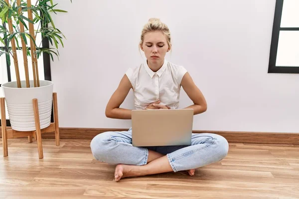 Young blonde woman using computer laptop sitting on the floor at the living room with hand on stomach because nausea, painful disease feeling unwell. ache concept.