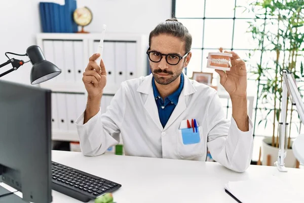Young hispanic dentist man holding denture and toothbrush skeptic and nervous, frowning upset because of problem. negative person. 