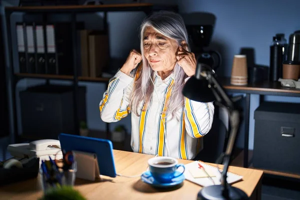 Middle age woman with grey hair working at the office at night covering ears with fingers with annoyed expression for the noise of loud music. deaf concept.
