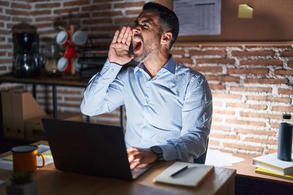 Hispanic man with beard working at the office at night shouting and screaming loud to side with hand on mouth. communication concept.