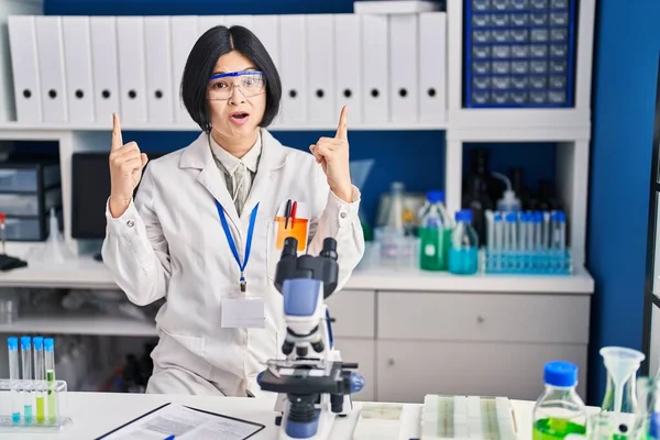 Young asian woman working at scientist laboratory amazed and surprised looking up and pointing with fingers and raised arms.