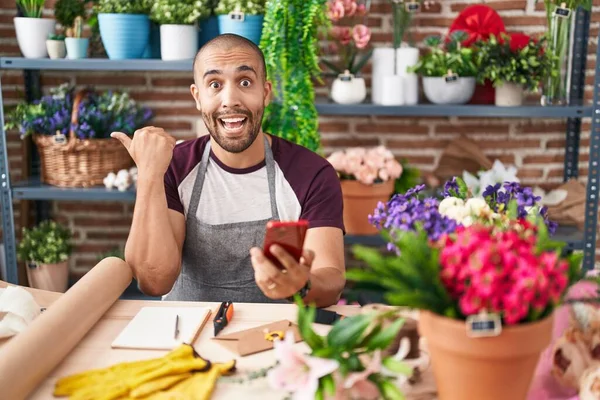 Hispanic man with beard working at florist shop with smartphone pointing thumb up to the side smiling happy with open mouth
