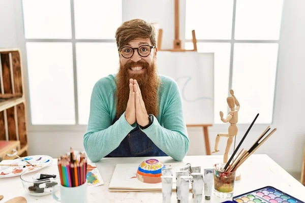 Redhead man with long beard painting clay bowl at art studio praying with hands together asking for forgiveness smiling confident.