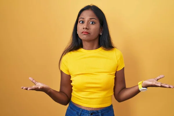 年轻的印度女人站在黄色的背景上 毫无头绪 与张开的双臂混淆不清 没有概念 — 图库照片