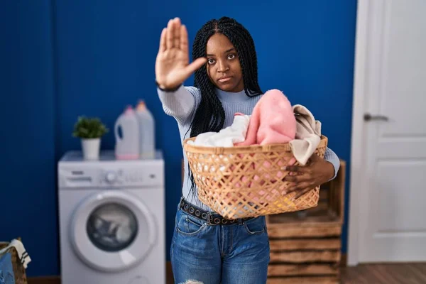 African american woman holding laundry basket with open hand doing stop sign with serious and confident expression, defense gesture