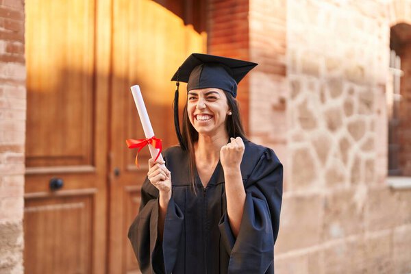 Young hispanic woman wearing graduated uniform holding diploma at university