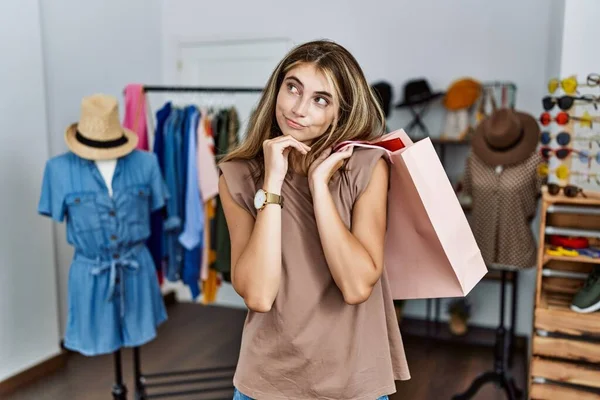 年轻的金发女人手托着购物袋在零售店里手托着下巴思考着问题 沉思着表情 脸上挂着沉思的笑容 怀疑概念 — 图库照片