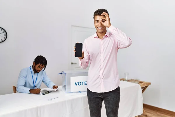 Young hispanic men at political campaign election holding smartphone smiling happy doing ok sign with hand on eye looking through fingers