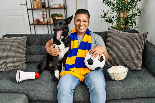 Young hispanic hooligan man supporting soccer team sitting on the sofa with dog at home.