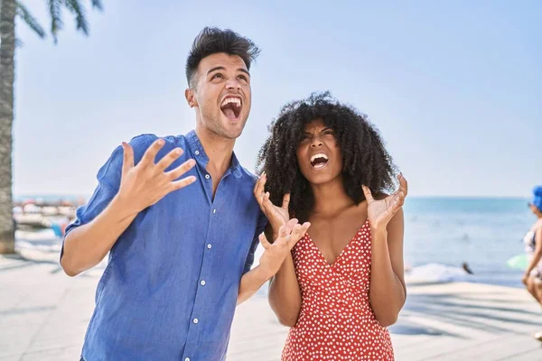 Young interracial couple outdoors on a sunny day crazy and mad shouting and yelling with aggressive expression and arms raised. frustration concept.