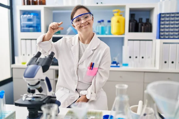 Hispanic girl with down syndrome working at scientist laboratory smiling and confident gesturing with hand doing small size sign with fingers looking and the camera. measure concept.