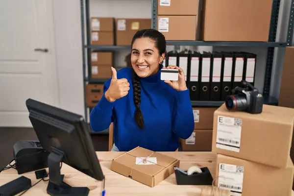 在小企业工作的年轻惊慌失措的女人拿着信用卡开心而积极地微笑着 大拇指向上做得出色 签了字 — 图库照片