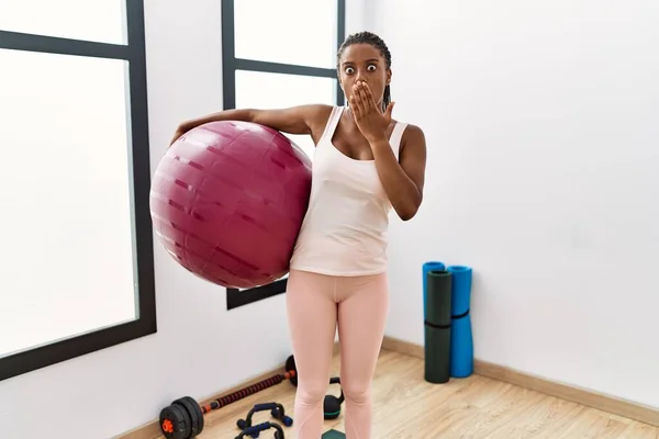 Young african american woman with braids holding pilate ball at yoga room covering mouth with hand, shocked and afraid for mistake. surprised expression