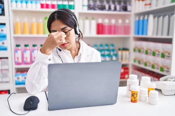 Young arab woman working at pharmacy drugstore using laptop tired rubbing nose and eyes feeling fatigue and headache. stress and frustration concept.