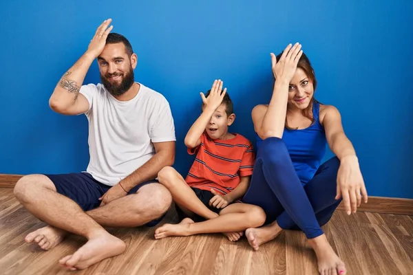Family of three sitting on the floor at home surprised with hand on head for mistake, remember error. forgot, bad memory concept.