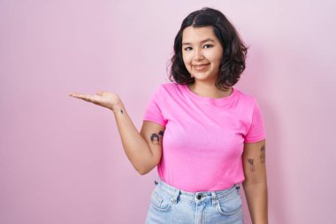 Young hispanic woman standing over pink background smiling cheerful presenting and pointing with palm of hand looking at the camera. 