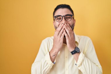 İş kıyafetleri ve gözlük takan İspanyol genç adam gülüyor ve utanıyor kıkır kıkır kıkır kıkır gülüyor eller, dedikodu ve skandal konseptiyle ağzını kapatıyor 